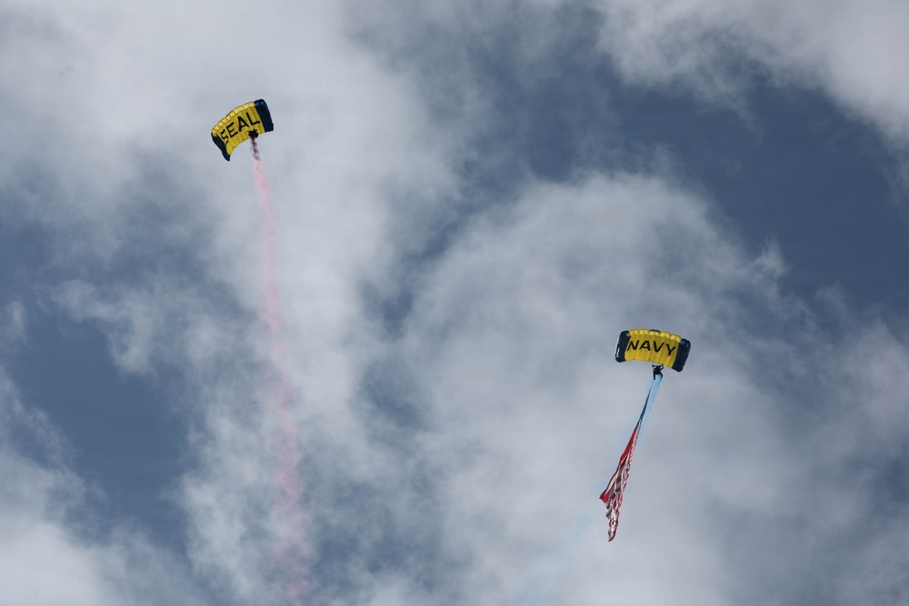 Two military skydivers are descending through a partly cloudy sky using yellow parachutes. One parachute is labeled 'SEAL' and is trailing red smoke, while the other is labeled 'NAVY' and carries what appears to be an American flag. This appears to be a ceremonial or demonstration parachute jump, likely performed by U.S. Navy SEALs.