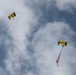 Two military skydivers are descending through a partly cloudy sky using yellow parachutes. One parachute is labeled 'SEAL' and is trailing red smoke, while the other is labeled 'NAVY' and carries what appears to be an American flag. This appears to be a ceremonial or demonstration parachute jump, likely performed by U.S. Navy SEALs.