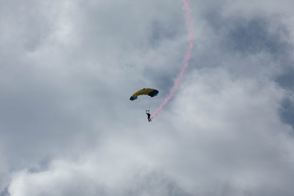 A parachutist with a yellow and dark-colored canopy is descending through an overcast sky. The skydiver is releasing a pink/red colored smoke trail that curves upward and to the right, indicating a recent spiral or turning maneuver. This appears to be a demonstration or airshow performance.