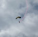 A parachutist with a yellow and dark-colored canopy is descending through an overcast sky. The skydiver is releasing a pink/red colored smoke trail that curves upward and to the right, indicating a recent spiral or turning maneuver. This appears to be a demonstration or airshow performance.