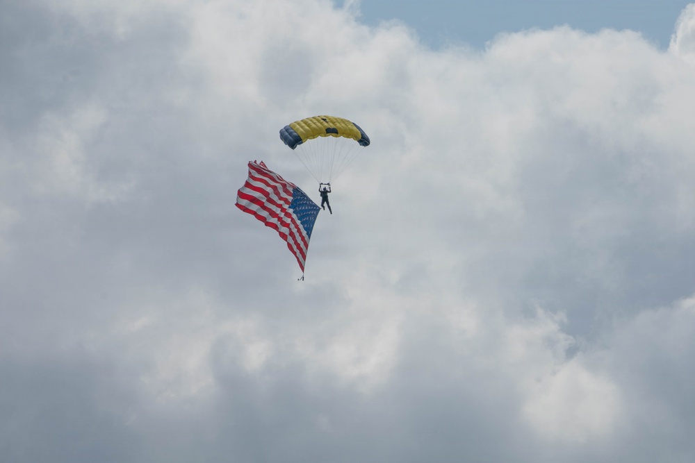 A skydiver descends under a yellow and dark blue parachute canopy while carrying a large American flag. The scene is set against a partly cloudy sky with white and grey cumulus clouds. This appears to be a ceremonial or demonstration skydive, likely at a public event or airshow.