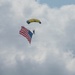 A skydiver descends under a yellow and dark blue parachute canopy while carrying a large American flag. The scene is set against a partly cloudy sky with white and grey cumulus clouds. This appears to be a ceremonial or demonstration skydive, likely at a public event or airshow.