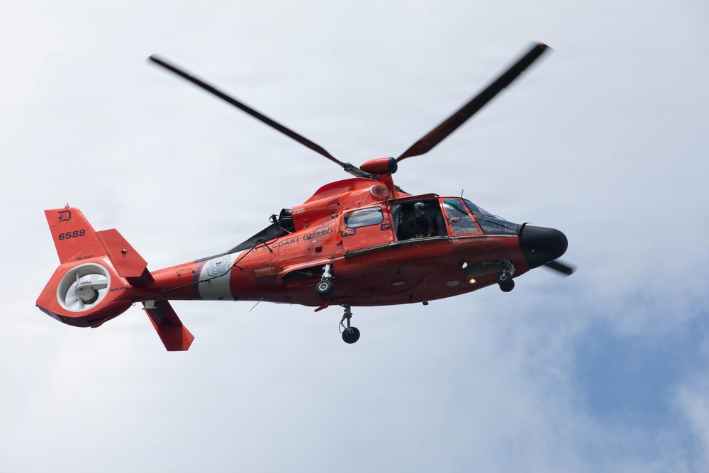 A United States Coast Guard helicopter, numbered 6588, is photographed in flight against an overcast sky. The aircraft is an MH-65 Dolphin helicopter, painted in the distinctive Coast Guard international orange color scheme. The helicopter appears to be hovering or in low-altitude flight, with rotor blades visibly in motion.