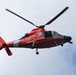 A United States Coast Guard helicopter, numbered 6588, is photographed in flight against an overcast sky. The aircraft is an MH-65 Dolphin helicopter, painted in the distinctive Coast Guard international orange color scheme. The helicopter appears to be hovering or in low-altitude flight, with rotor blades visibly in motion.