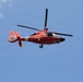 A bright orange U.S. Coast Guard helicopter is captured in flight against a clear blue sky with light cloud cover. The aircraft appears to be a Eurocopter MH-65 Dolphin, identifiable by its distinctive orange livery and fenestron (enclosed tail rotor). The tail number '6588' is visible on the tail boom.