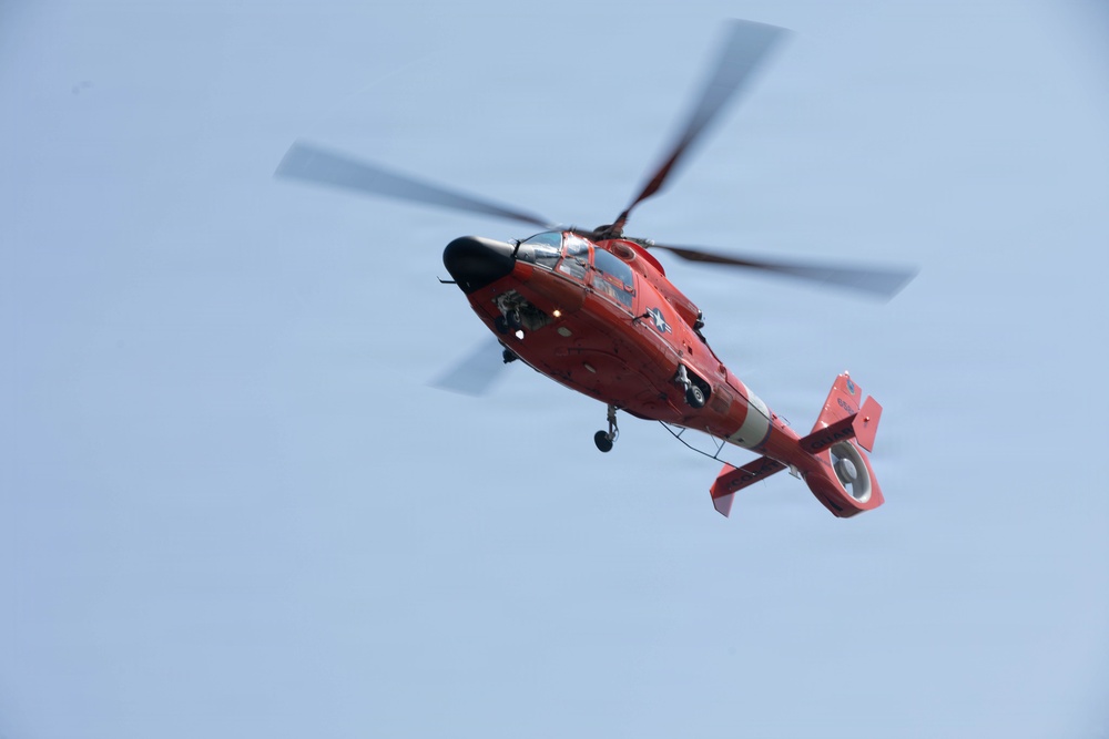 A red and white helicopter is captured in flight against a clear light blue sky. The helicopter displays markings consistent with a United States Coast Guard MH-65 Dolphin rescue helicopter. The main rotor blades are visibly motion-blurred, indicating active flight.