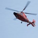 A red and white helicopter is captured in flight against a clear light blue sky. The helicopter displays markings consistent with a United States Coast Guard MH-65 Dolphin rescue helicopter. The main rotor blades are visibly motion-blurred, indicating active flight.