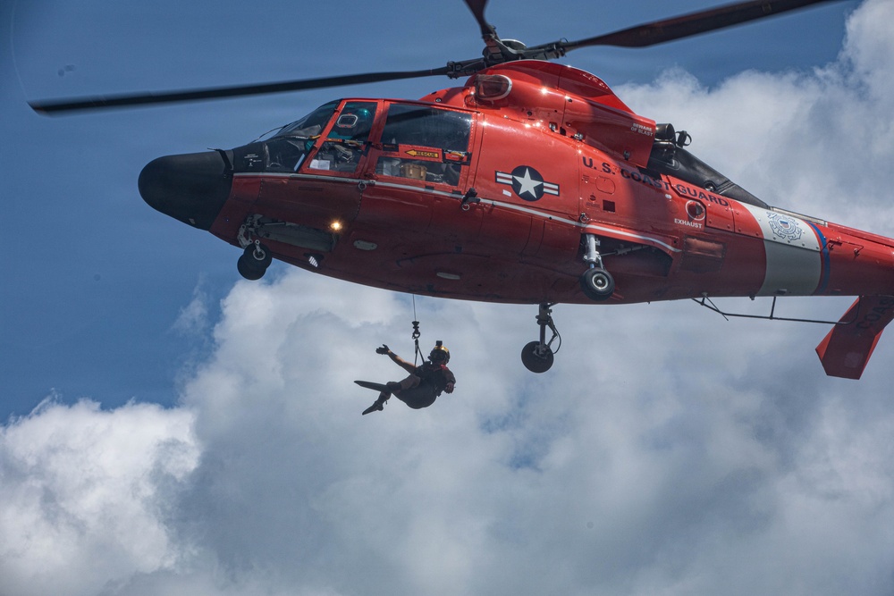 A U.S. Coast Guard helicopter, painted in its distinctive international orange color, is shown hovering while a rescue swimmer or crew member is suspended below on a hoist cable. The aircraft displays the U.S. Coast Guard insignia and a military star emblem on its fuselage. This appears to be either a rescue operation or a training exercise involving hoist deployment.