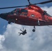 A U.S. Coast Guard helicopter, painted in its distinctive international orange color, is shown hovering while a rescue swimmer or crew member is suspended below on a hoist cable. The aircraft displays the U.S. Coast Guard insignia and a military star emblem on its fuselage. This appears to be either a rescue operation or a training exercise involving hoist deployment.