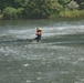 A person is being pulled across a fast-moving river or waterway while suspended from a zip line or cable system overhead. They are wearing safety gear including a helmet and life vest, suggesting this is either a rescue operation or a controlled adventure activity. The water below is turbulent and churning, indicating strong current.