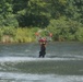 A rescue worker in dark clothing and a helmet stands waist-deep in a fast-moving river, holding onto a metal rescue basket suspended from an overhead cable system. The basket appears to contain red equipment or gear and is fitted with red flotation devices. The scene is set against a lush green tree line, suggesting a swift-water rescue operation or training exercise.