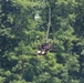A person is zip-lining through a dense, green forested area, suspended from a cable above. The individual is wearing safety gear including a harness, helmet, and goggles, and appears to be in a reclined position mid-flight. The lush green tree canopy serves as the backdrop, suggesting a warm-season outdoor adventure activity.