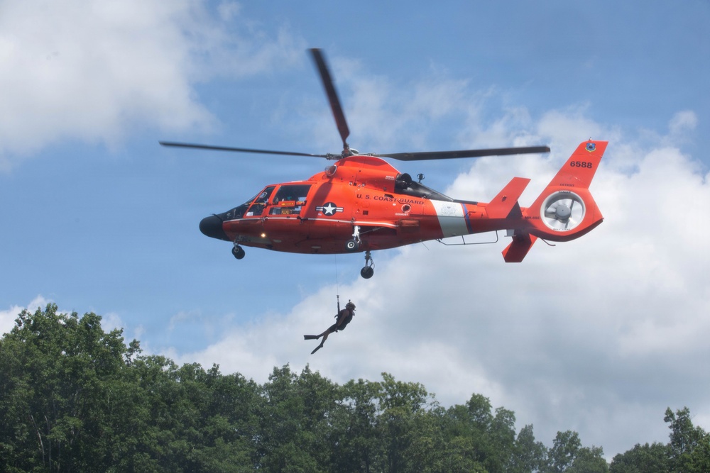 A U.S. Coast Guard helicopter (tail number 6588) hovers in the air while a rescue swimmer is being lowered on a hoist cable. The bright orange MH-65 Dolphin helicopter displays the U.S. Coast Guard insignia and a military star emblem on its fuselage. The scene appears to be a training exercise or demonstration conducted over a wooded area.