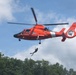 A U.S. Coast Guard helicopter (tail number 6588) hovers in the air while a rescue swimmer is being lowered on a hoist cable. The bright orange MH-65 Dolphin helicopter displays the U.S. Coast Guard insignia and a military star emblem on its fuselage. The scene appears to be a training exercise or demonstration conducted over a wooded area.