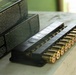 The image shows a row of brass-cased ammunition cartridges arranged in a black plastic ammunition tray or holder on a light-colored surface. Two dark-colored ammunition boxes are visible in the background to the left. The setting appears to be an outdoor or semi-outdoor area, possibly a shooting range.