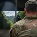 Two U.S. Army soldiers in OCP (Operational Camouflage Pattern) uniforms observe a shooting range activity from a covered firing position. The soldier in the foreground has a name tape reading 'HALL' on his patrol cap. A shooter in civilian/dark clothing is visible in the background actively firing downrange toward a wooded hillside.