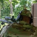 A person named 'Paul' (as labeled on their cap) is positioned at a shooting range bench, aiming a bolt-action or precision rifle equipped with a large magnification scope and bipod. The individual is wearing multicam/OCP camouflage clothing and electronic hearing protection earmuffs. The setting appears to be an outdoor shooting range with dense green vegetation visible downrange.