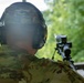 A military or law enforcement personnel identified as 'PAUL' is positioned at a shooting range, facing downrange with a scoped rifle on a bipod mounted on a surface in front of them. The individual is wearing multicam/OCP pattern camouflage uniform, a matching helmet, and electronic hearing protection (Impact Sport earmuffs). A two-way radio is visible on the right side of the shooting bench.