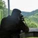 A person wearing camouflage military-style clothing and a cap is positioned at a wooden shooting bench, aiming a scoped rifle mounted on a bipod toward a forested hillside. The setting appears to be an outdoor shooting range with a covered firing position. A detached magazine is visible on the wooden bench surface to the right.
