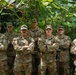 Seven U.S. Army soldiers in MultiCam/OCP camouflage uniforms pose together for a group photo outdoors in a wooded or heavily vegetated area. All soldiers have their arms crossed and are facing the camera, suggesting a formal team or unit photograph. Name tapes and U.S. Army tapes are visible on their uniforms, along with various rank insignia and shoulder patches.