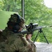 A person in military camouflage uniform is firing a precision rifle at an outdoor shooting range. A spent brass casing is visibly ejecting from the rifle's ejection port, captured mid-air. The shooter is positioned at a wooden bench with a forested valley visible in the background.