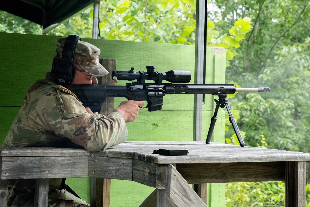A person wearing military camouflage (OCP/MultiCam pattern) uniform and cap is positioned at a wooden shooting bench, aiming a precision bolt-action rifle equipped with a scope and bipod. The setting appears to be an outdoor covered shooting range with green-painted wooden structures and dense foliage visible in the background. The shooter is wearing hearing protection ear muffs and appears to be in a focused firing position.