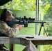 A person wearing military camouflage (OCP/MultiCam pattern) uniform and cap is positioned at a wooden shooting bench, aiming a precision bolt-action rifle equipped with a scope and bipod. The setting appears to be an outdoor covered shooting range with green-painted wooden structures and dense foliage visible in the background. The shooter is wearing hearing protection ear muffs and appears to be in a focused firing position.