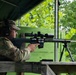 A soldier in military camouflage uniform is positioned at an outdoor shooting range, aiming a precision bolt-action rifle mounted on a bipod. The individual is wearing hearing protection labeled 'Impact Sport' and eye protection while in a prone-ready firing position at a wooden bench. The setting appears to be a covered range facility surrounded by dense green foliage.