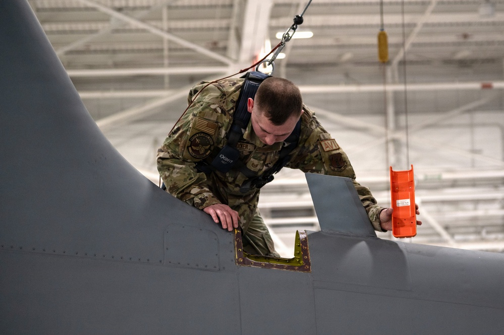 A U.S. Air Force maintainer in OCP (Operational Camouflage Pattern) uniform is performing maintenance or inspection on top of a military aircraft inside a hangar. The airman is wearing a safety harness connected to an overhead rigging system and is holding an orange tool or component while examining a panel on the aircraft's fuselage. An open access panel with visible internal structure is present on the aircraft surface.
