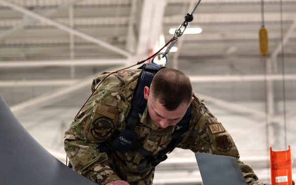 A U.S. Air Force maintainer in OCP (Operational Camouflage Pattern) uniform is performing maintenance or inspection on top of a military aircraft inside a hangar. The airman is wearing a safety harness connected to an overhead rigging system and is holding an orange tool or component while examining a panel on the aircraft's fuselage. An open access panel with visible internal structure is present on the aircraft surface.