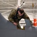 A U.S. Air Force maintainer in OCP (Operational Camouflage Pattern) uniform is performing maintenance or inspection on top of a military aircraft inside a hangar. The airman is wearing a safety harness connected to an overhead rigging system and is holding an orange tool or component while examining a panel on the aircraft's fuselage. An open access panel with visible internal structure is present on the aircraft surface.