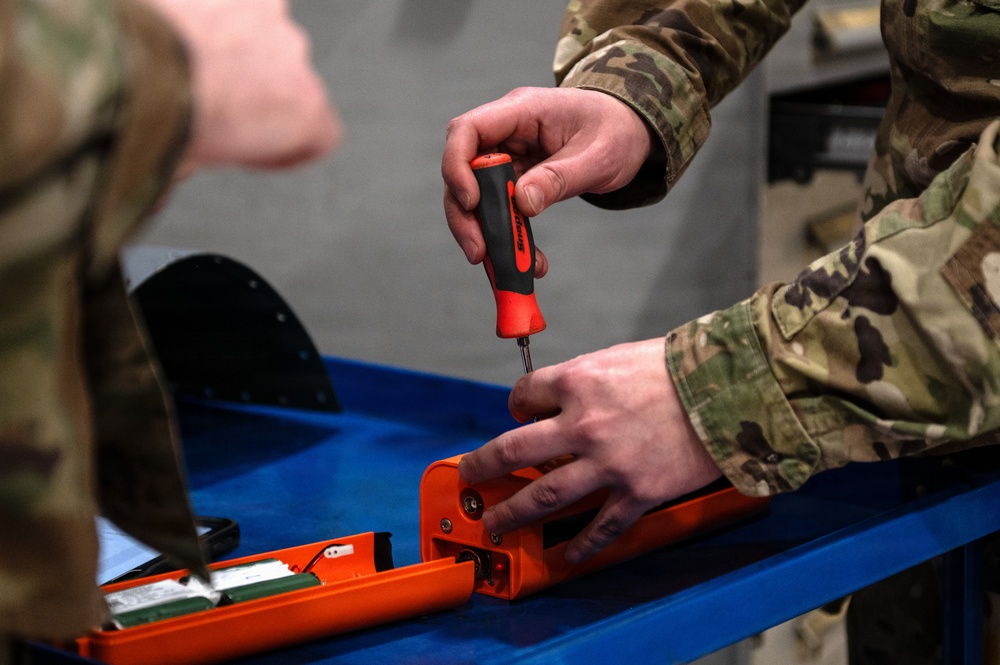 A military service member in MultiCam/OCP camouflage uniform is using a red and black screwdriver to work on an orange-colored device on a blue workbench. The orange device appears to be an electronic or mechanical component, possibly a flight data recorder, emergency locator transmitter, or similar equipment. A second uniformed individual is partially visible in the background.