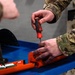 A military service member in MultiCam/OCP camouflage uniform is using a red and black screwdriver to work on an orange-colored device on a blue workbench. The orange device appears to be an electronic or mechanical component, possibly a flight data recorder, emergency locator transmitter, or similar equipment. A second uniformed individual is partially visible in the background.