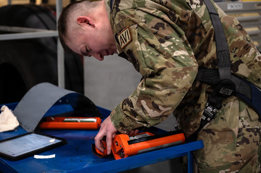 A military service member wearing OCP (Operational Camouflage Pattern) uniform with an 'AVI' name tape is leaning over a blue work cart, handling an orange-colored tool or device. The individual is also wearing a safety harness, suggesting work in an elevated or hazardous environment. The setting appears to be a military maintenance or hangar facility.