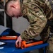 A military service member wearing OCP (Operational Camouflage Pattern) uniform with an 'AVI' name tape is leaning over a blue work cart, handling an orange-colored tool or device. The individual is also wearing a safety harness, suggesting work in an elevated or hazardous environment. The setting appears to be a military maintenance or hangar facility.