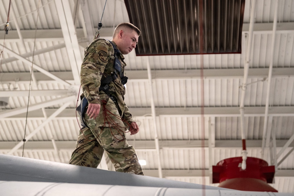 A military service member in OCP (Operational Camouflage Pattern) uniform is walking on top of a large white aircraft wing or fuselage inside a hangar. The individual is wearing a safety harness connected to an overhead fall-arrest system, suggesting this is a maintenance or inspection activity. A red-capped component, likely a munition or fuel tank, is visible in the background.