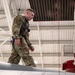 A military service member in OCP (Operational Camouflage Pattern) uniform is walking on top of a large white aircraft wing or fuselage inside a hangar. The individual is wearing a safety harness connected to an overhead fall-arrest system, suggesting this is a maintenance or inspection activity. A red-capped component, likely a munition or fuel tank, is visible in the background.