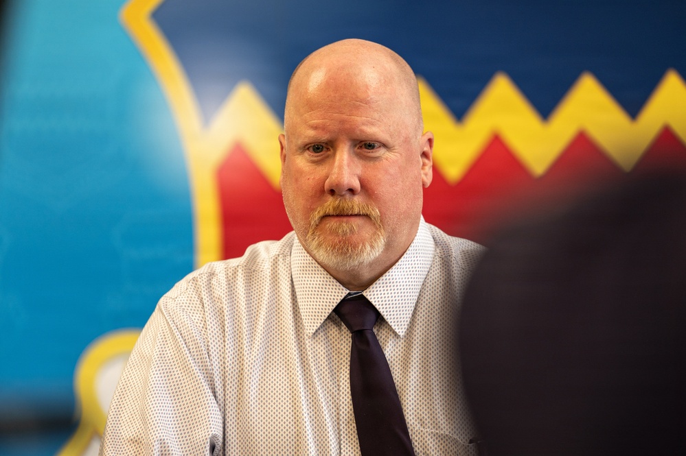 A bald middle-aged man with a goatee is seated in what appears to be a professional or institutional setting. He is wearing a white patterned dress shirt and a dark burgundy/maroon tie. Behind him is a colorful mural or wall decoration featuring blue, red, and yellow zigzag patterns.