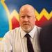 A bald middle-aged man with a goatee is seated in what appears to be a professional or institutional setting. He is wearing a white patterned dress shirt and a dark burgundy/maroon tie. Behind him is a colorful mural or wall decoration featuring blue, red, and yellow zigzag patterns.