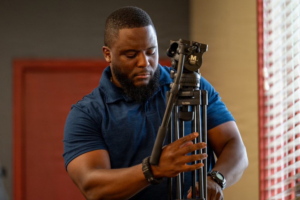 A man wearing a blue polo shirt is handling a professional video tripod, appearing to adjust or set it up. He has a beard, short hair, and is wearing a watch on his left wrist. The setting appears to be an indoor environment with a red door and window blinds visible in the background.