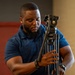 A man wearing a blue polo shirt is handling a professional video tripod, appearing to adjust or set it up. He has a beard, short hair, and is wearing a watch on his left wrist. The setting appears to be an indoor environment with a red door and window blinds visible in the background.