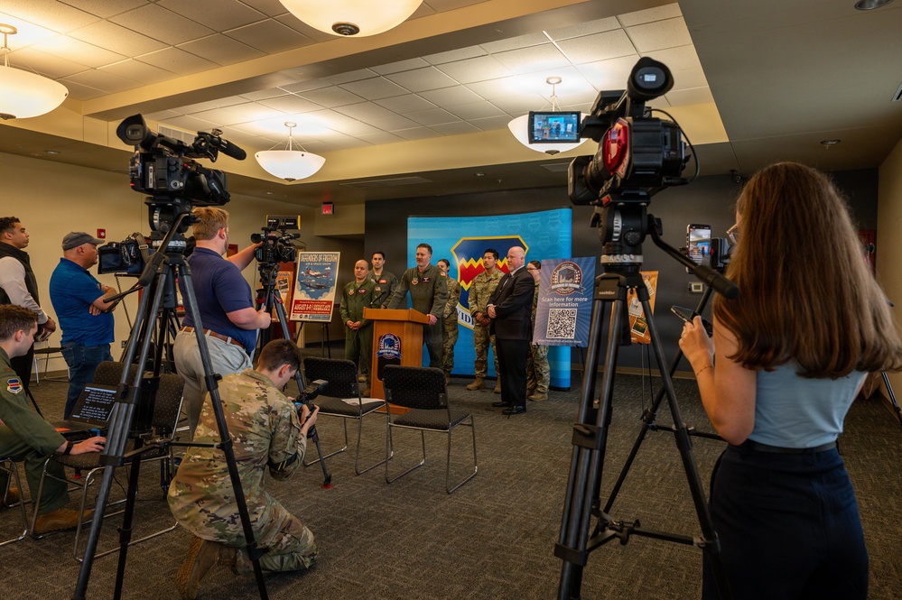 A military press conference is being held in an indoor briefing room, with several uniformed military personnel and at least one civilian standing at a podium in front of a blue backdrop bearing a military unit insignia. Multiple camera operators and media personnel are filming and photographing the event. Promotional banners and posters, including one with a QR code, are visible flanking the speakers.
