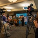 A military press conference is being held in an indoor briefing room, with several uniformed military personnel and at least one civilian standing at a podium in front of a blue backdrop bearing a military unit insignia. Multiple camera operators and media personnel are filming and photographing the event. Promotional banners and posters, including one with a QR code, are visible flanking the speakers.