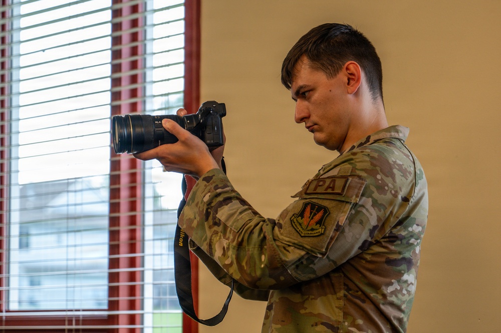 A young military service member in OCP (Operational Camouflage Pattern) uniform is holding and operating a Nikon DSLR camera with a large telephoto or zoom lens. The individual wears a 'PA' (Public Affairs) patch on their chest and a unit patch on their left shoulder, indicating they are a military public affairs photographer. The photo is taken indoors near a window with horizontal blinds.