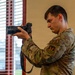 A young military service member in OCP (Operational Camouflage Pattern) uniform is holding and operating a Nikon DSLR camera with a large telephoto or zoom lens. The individual wears a 'PA' (Public Affairs) patch on their chest and a unit patch on their left shoulder, indicating they are a military public affairs photographer. The photo is taken indoors near a window with horizontal blinds.