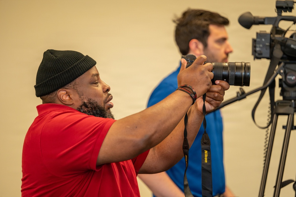 Two men are engaged in photography and videography in what appears to be an indoor studio or event setting with a neutral beige backdrop. The man in the foreground, wearing a red shirt and black beanie, is actively shooting with a Nikon DSLR camera with a zoom lens. A second man in a blue shirt is positioned behind a professional video camera mounted on a tripod.