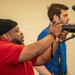 Two men are engaged in photography and videography in what appears to be an indoor studio or event setting with a neutral beige backdrop. The man in the foreground, wearing a red shirt and black beanie, is actively shooting with a Nikon DSLR camera with a zoom lens. A second man in a blue shirt is positioned behind a professional video camera mounted on a tripod.
