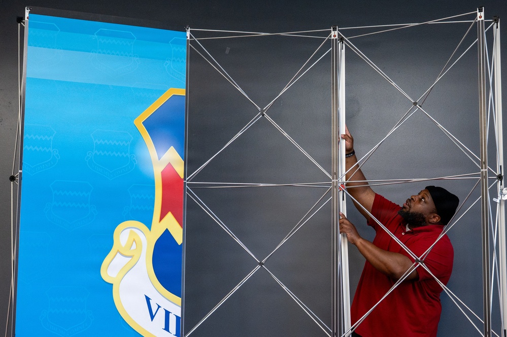 A man wearing a red polo shirt and dark cap is assembling a large pop-up display stand or trade show backdrop frame. The collapsible aluminum lattice framework is partially erected, with one panel already covered by a blue graphic banner featuring a shield/crest logo with the partial text 'VI'. The setting appears to be an indoor venue, likely being prepared for an event or exhibition.