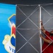 A man wearing a red polo shirt and dark cap is assembling a large pop-up display stand or trade show backdrop frame. The collapsible aluminum lattice framework is partially erected, with one panel already covered by a blue graphic banner featuring a shield/crest logo with the partial text 'VI'. The setting appears to be an indoor venue, likely being prepared for an event or exhibition.