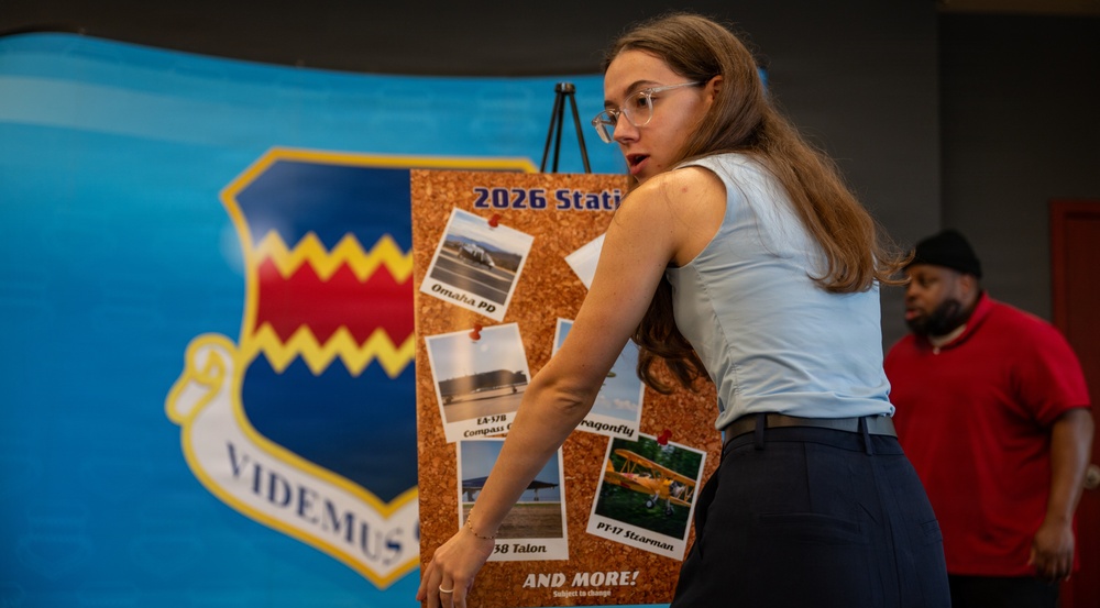 A young woman with glasses and long brown hair is presenting or pointing to a cork bulletin board displaying photos and information about a '2026 Static' display event. The board features photographs of various aircraft including an EA-37B, Dragonfly, T-38 Talon, and PT-17 Stearman, along with a reference to 'Omaha PD.' Behind her is a large blue banner featuring a military unit shield with the motto 'VIDEMUS.'